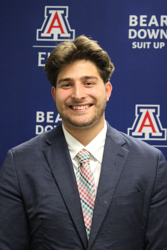Portrait of Blake Raben, smiling, in a navy suit and checked tie in front of a University of Arizona backdrop